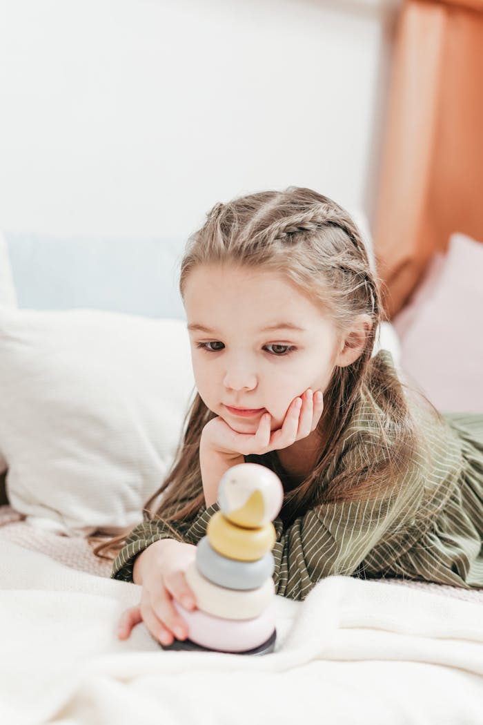 A young girl with braids playing with a colorful stacking toy indoors, focusing intently and showing curiosity.