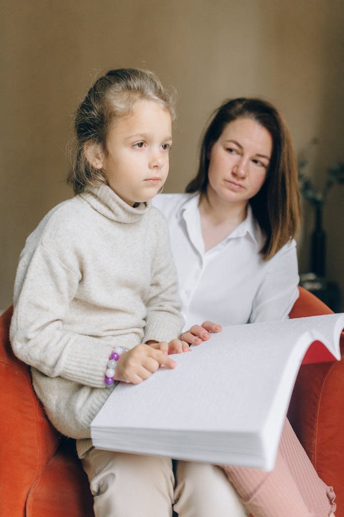 A mother supports her daughter as she reads braille indoors, fostering learning and bonding.